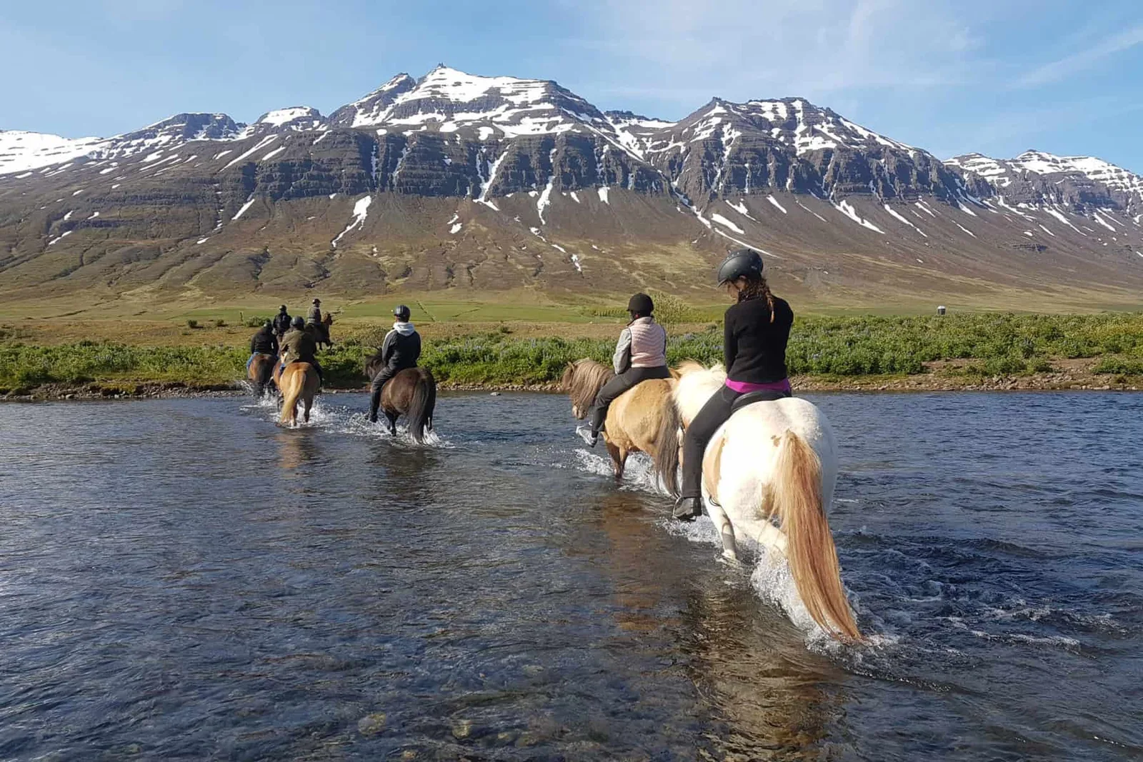 Reiðfólk fer yfir á á íslenskum hestum með snævi þakin fjöll í baksýn.